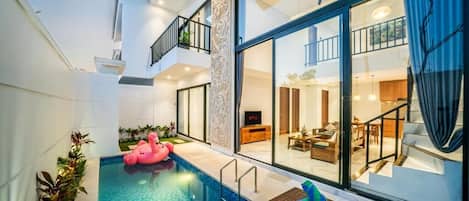 A modern indoor pool area with a glass wall, a floating pink flamingo, and a wicker lounge chair.