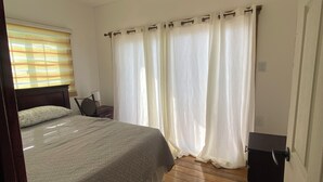 A bedroom with a bed, wooden floor, curtains, and a nightstand.