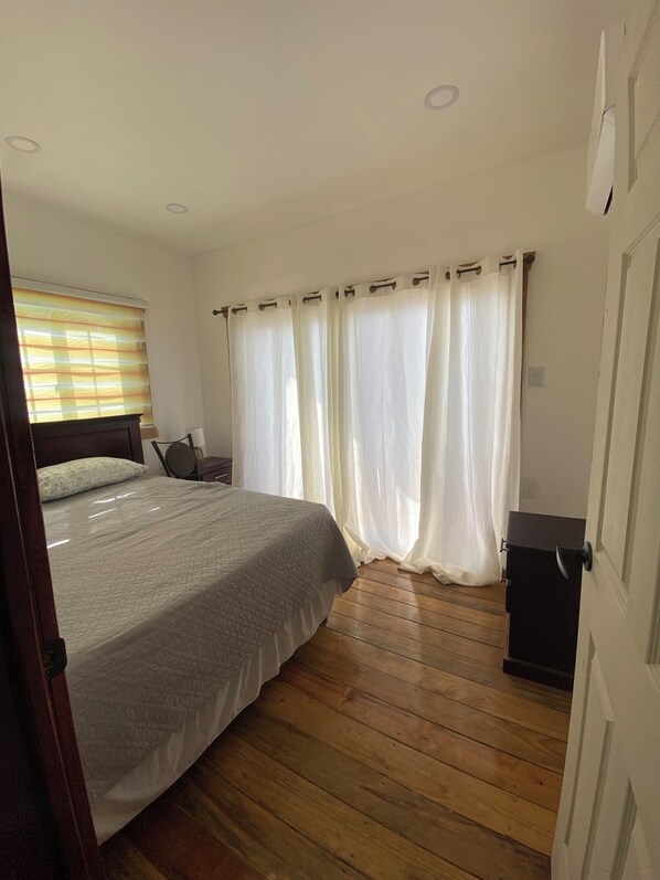 A bedroom with a bed, wooden flooring, a nightstand, and a window with blinds. - Ocean Breeze Villas (Sambo Creek)