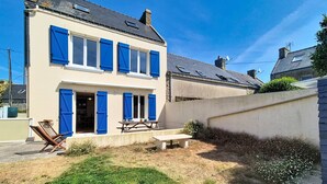 Een tweelaags huis met blauwe luiken, een terras met tafel en stoelen, en een verzorgd gazon.