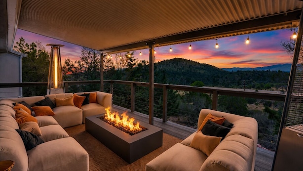 Ein überdachter Außenbereich mit Sitzgelegenheit, einem Feuerstelle, einer Couch und Blick auf einen Sonnenuntergang über einer hügeligen Landschaft.