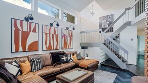 Ein modernes Wohnzimmer mit einem braunen Ledersofa, abstrakter Wandkunst, einer Treppe und Blick auf den Himmel.