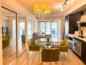 A modern kitchen with stainless steel appliances, a dining area with a glass table and green chairs, and a large window offering a city view. - West Queen Atelier (Toronto)
