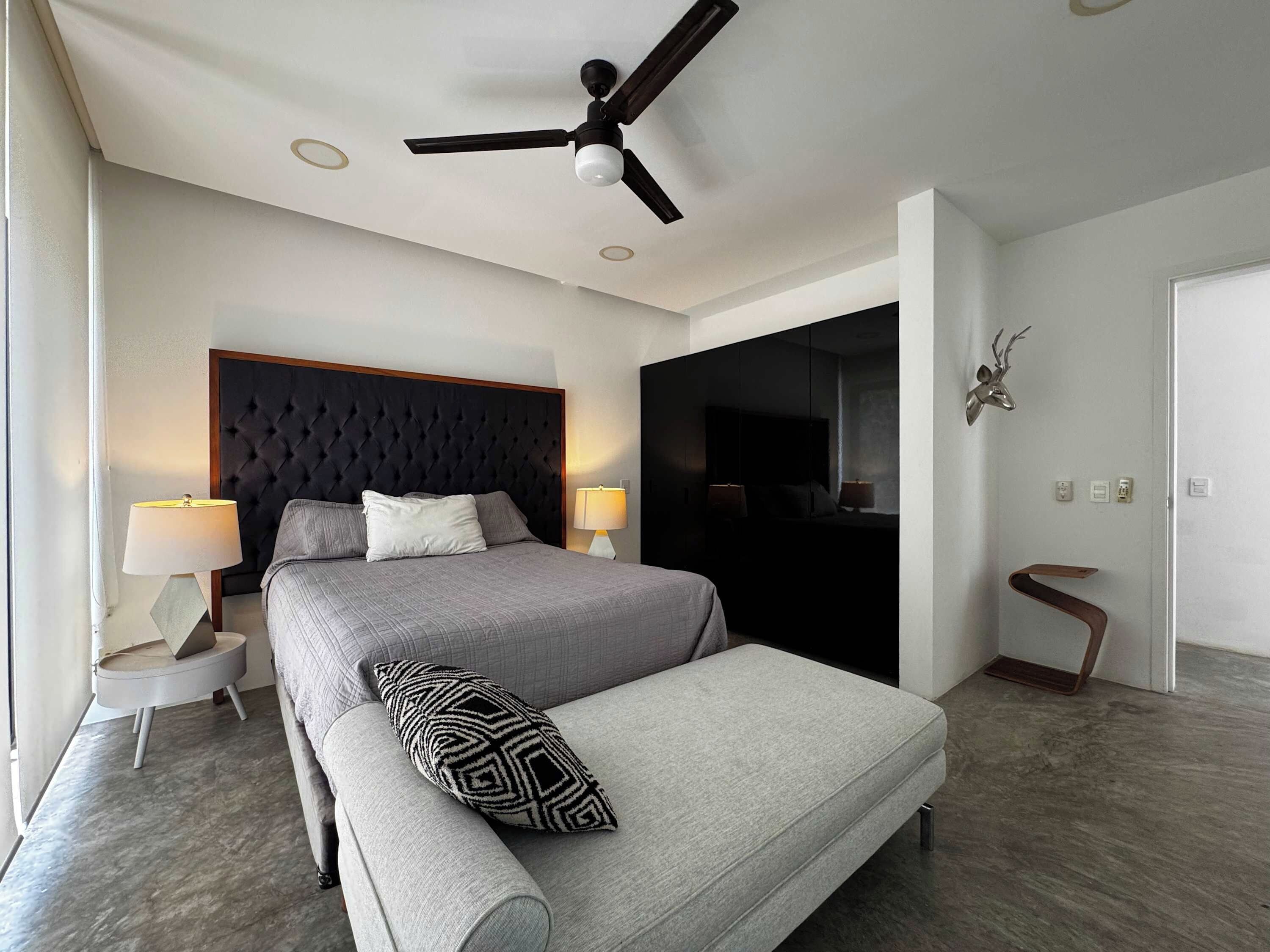 A modern hotel room with a bed, two bedside lamps, a ceiling fan, a large mirror, and a deer head decoration.