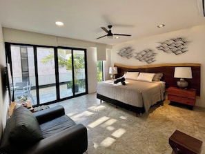 A hotel room with a bed, a nightstand, a chair, a ceiling fan, and a view through the sliding glass doors. - Black House - Stylish 3BR in Aldea Zama with Rooftop (Tulum)