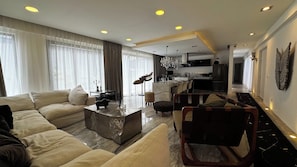 A modern living room with a sofa, coffee table, and ottoman, a dining area with chairs, and a kitchen in the background. - Black House - Stylish 3BR in Aldea Zama with Rooftop (Tulum)