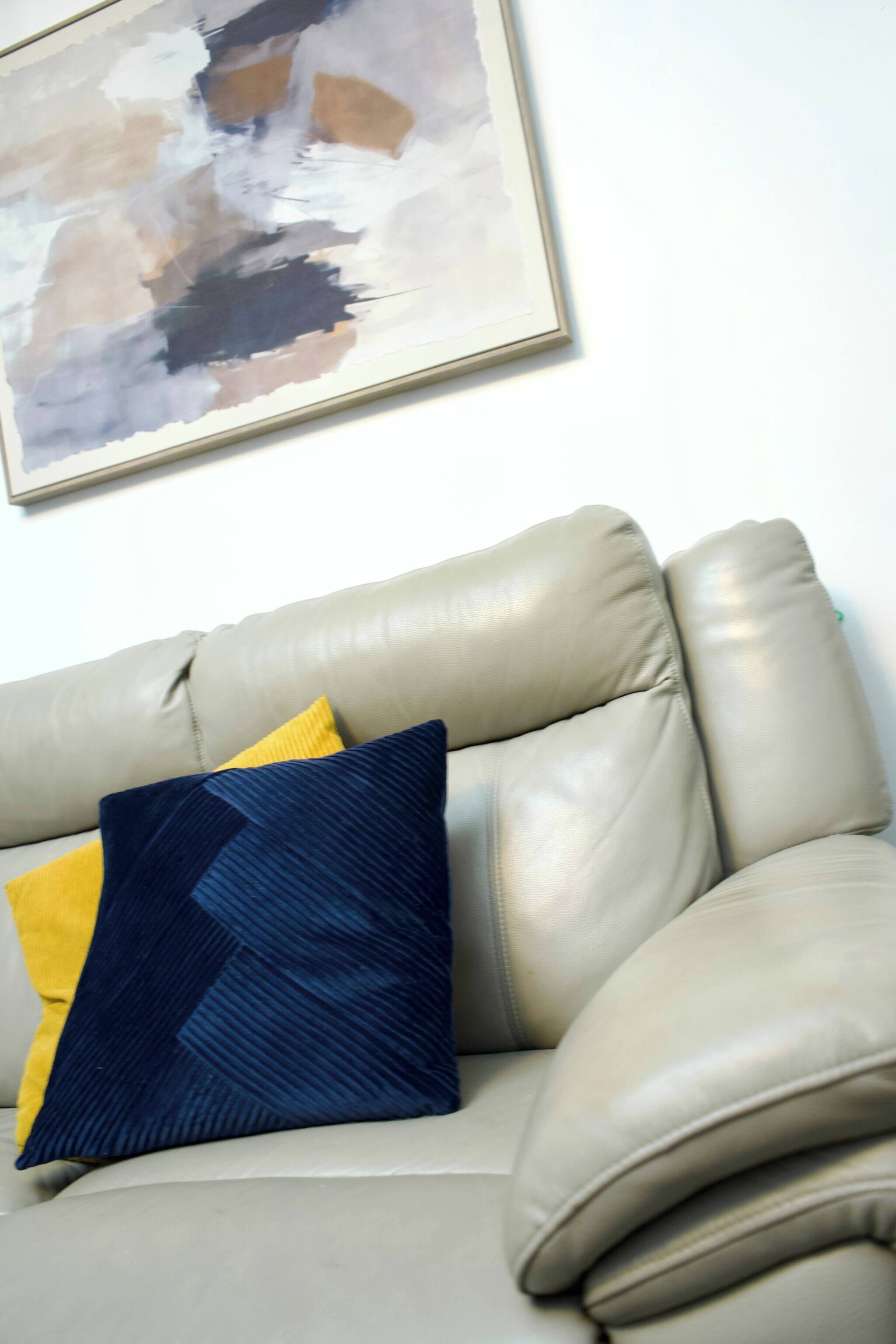 Ein beiges Ledersofa mit zwei Kissen, einem gelben und einem dunkelblauen, vor einer weißen Wand mit einem gerahmten abstrakten Gemälde.