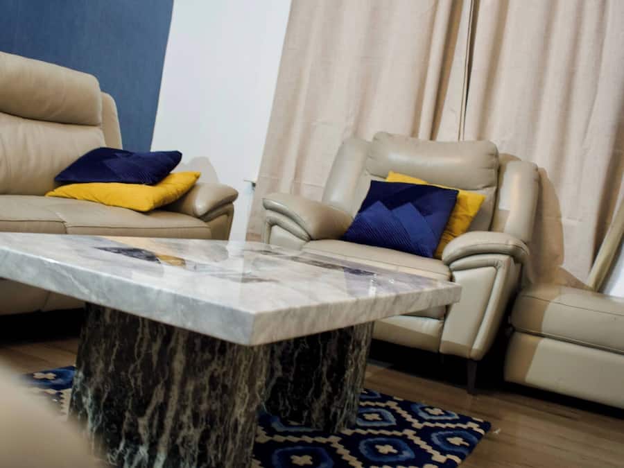 Ein Wohnzimmer mit einem beigen Sofa, einem Liegesessel, einem мраморному журнальному Tisch und einem gemusterten Teppich.