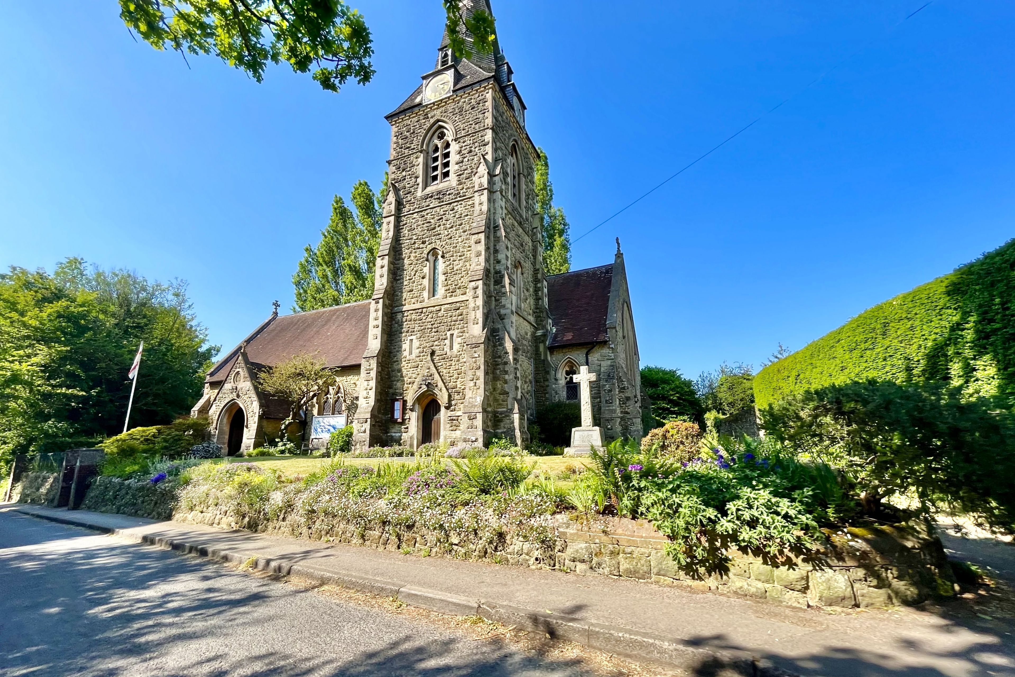 Une église en pierre, dotée d’un haut clocher et de fenêtres en arc de cercle, entourée d’une végétation luxuriante et surplombée d’un ciel bleu clair.