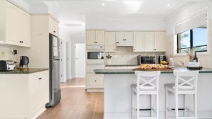 A modern kitchen with white cabinetry, a stainless steel refrigerator, a microwave, and a breakfast bar with two white chairs.