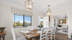 A dining area with a wooden table, white chairs, and a view through large windows.
