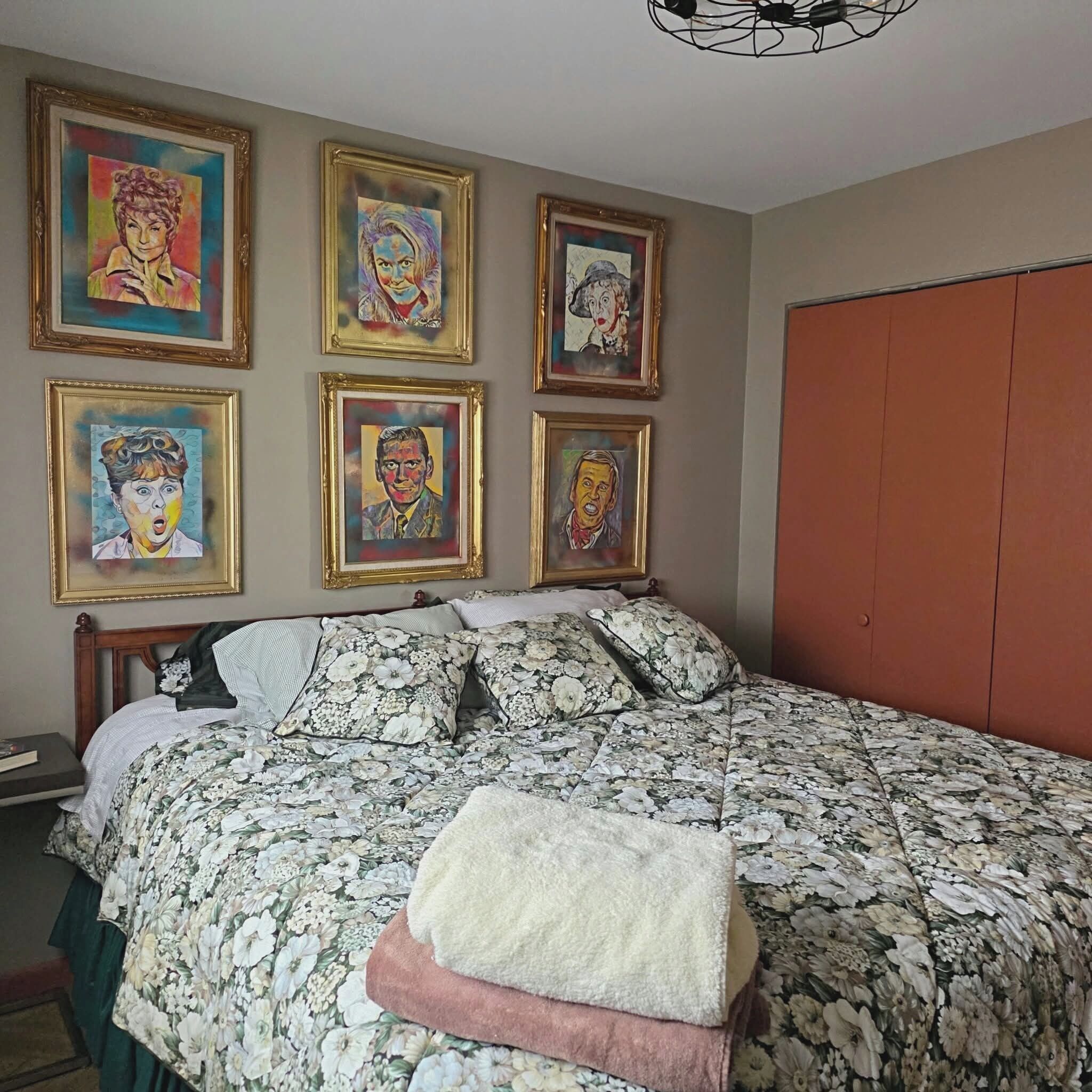 A bedroom with a floral bedspread, a wooden headboard, and six framed artworks on the wall.