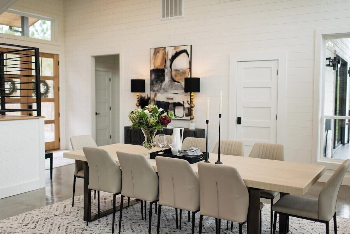 A dining area with a long table, six chairs, a vase with flowers, two wall-mounted lamps, and a large abstract painting.