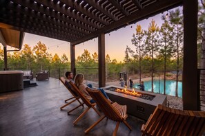 A covered outdoor seating area with a fire feature and a view of a swimming pool. - NEW! Pondside Lux Cabin with Hot Tub, King Suites, Fire Pit & Game Loft (Broken Bow)