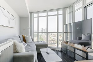 A modern living room with a sectional sofa, a flat-screen TV, and large windows offering a city view. - King West Place (Toronto)