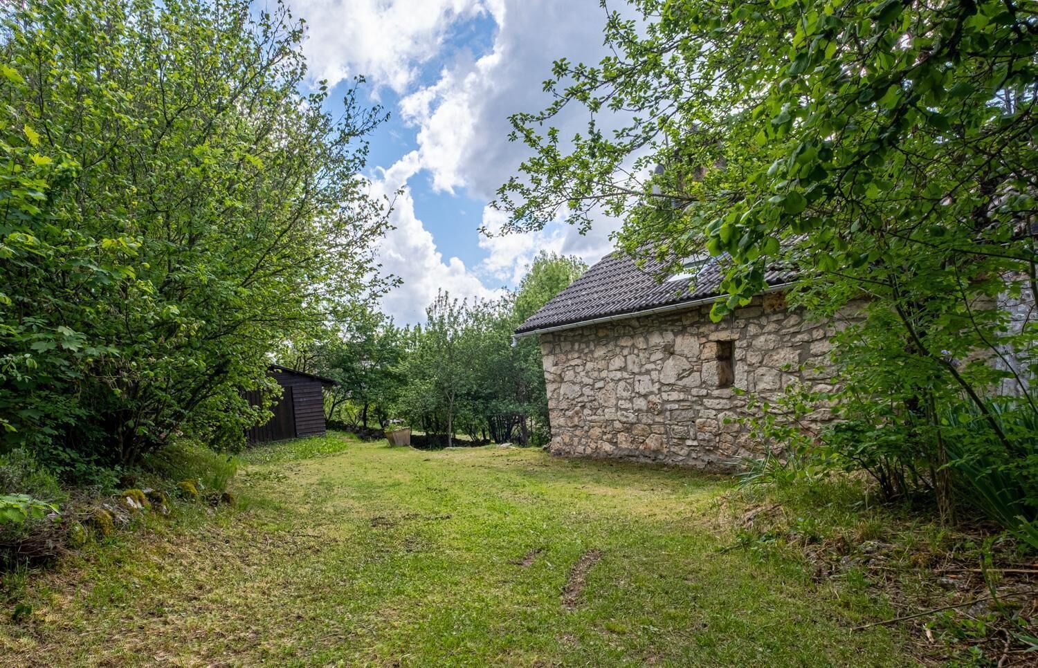 Un bâtiment en pierre, surmonté d’un toit de tuiles, entouré d’une végétation luxuriante et d’arbres.