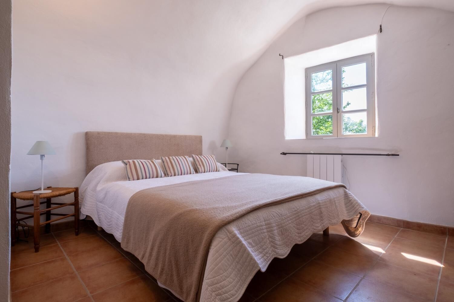 A bedroom with a bed, a nightstand, a lamp, a window, and a radiator.