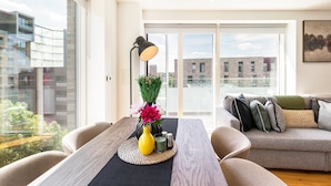 Ein modernes Wohnzimmer mit einer Couch, einem Tisch mit Blumenvase, einer Stehlampe und großen Fenstern mit Blick auf die Stadt.