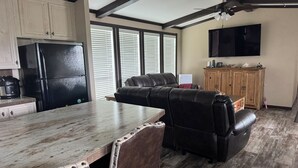 A kitchen with a black refrigerator, wooden cabinets, a dining table with a rustic design, a leather sofa, and a television mounted on the wall.
