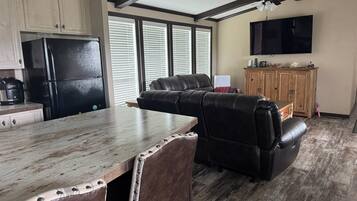 A kitchen with a black refrigerator, wooden cabinets, a dining table with a rustic design, a leather sofa, and a television mounted on the wall.