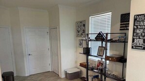 Un interior de hogar con piso de baldosas, una puerta blanca, un estante negro con diversos objetos y una pared con un cartel de normas familiares.