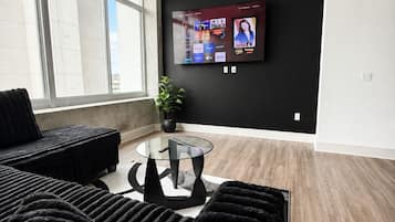 A modern living room with a large TV mounted on the wall, a ceiling fan, and a large window providing natural light.