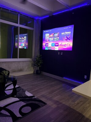 A room with a large flat-screen TV mounted on a dark wall, a window with blue LED lights, a potted plant, and a wooden floor.