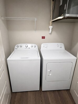 A laundry room with two white Whirlpool washers and dryers, a white shelf above, and a red safety sign on the wall.