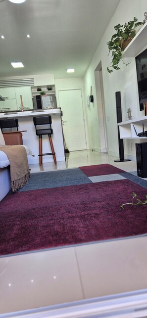 Een moderne woonkamer met een bank, een salontafel, een rood vloerkleed, een keuken met een aanrecht en barkrukken, en een plant op een plank.