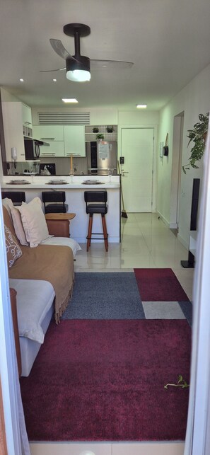 Een moderne woonkamer met een bank, een salontafel en een televisie. Er is een keukenhoek met een koelkast, magnetron en een eettafel met stoelen.