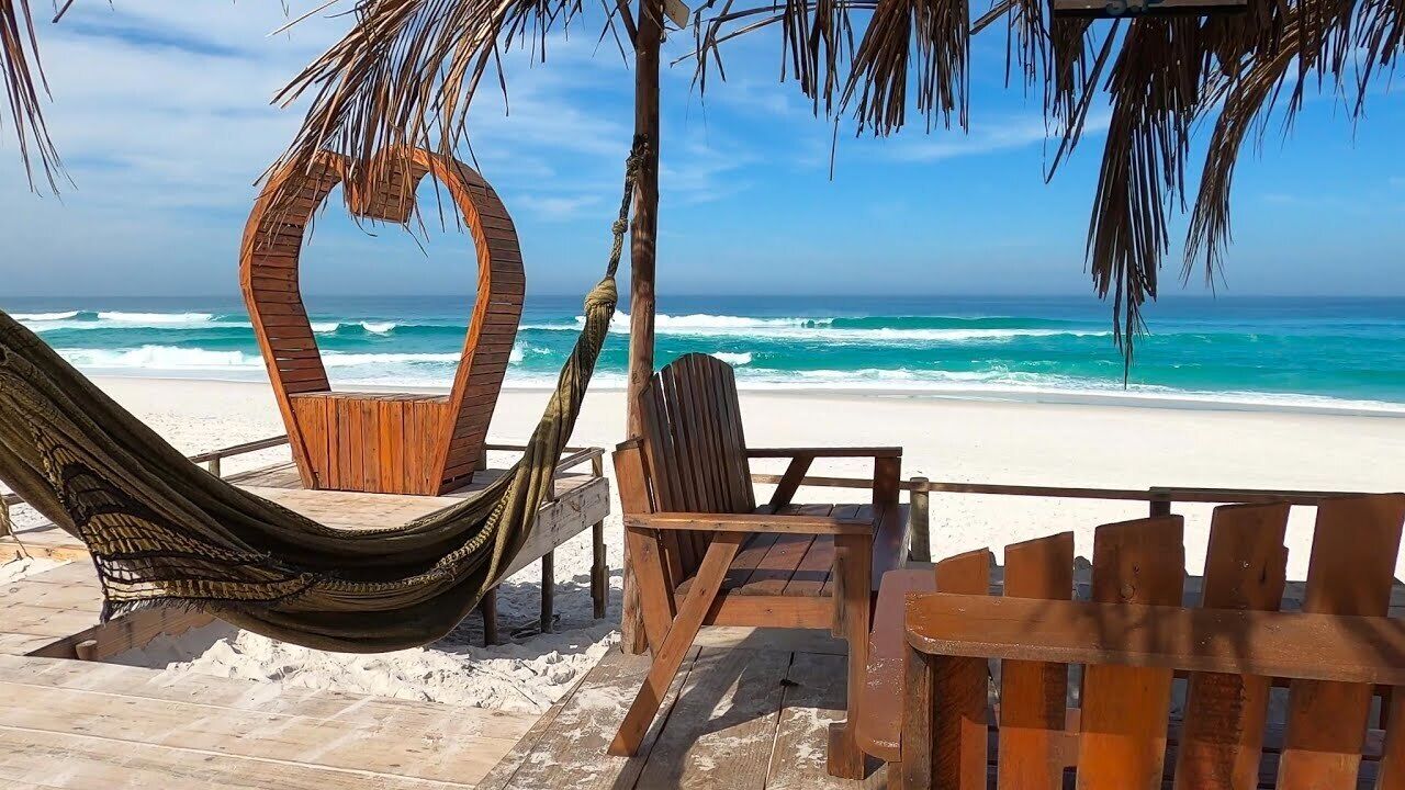 Une terrasse en bois avec un hamac, une chaise en bois et une table en bois, donnant sur une plage où l’on peut admirer les vagues.