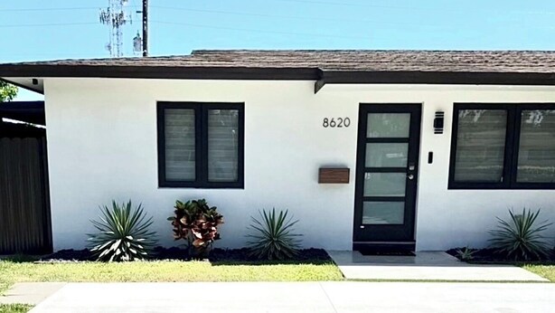 Une maison moderne à deux étages, avec le numéro 8620 sur la porte, une pelouse et des plantes devant, et un ciel dégagé.