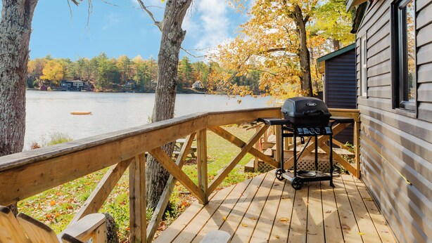 Una terraza de madera con una parrilla, con vista a un lago rodeado de árboles y una cabaña al fondo.