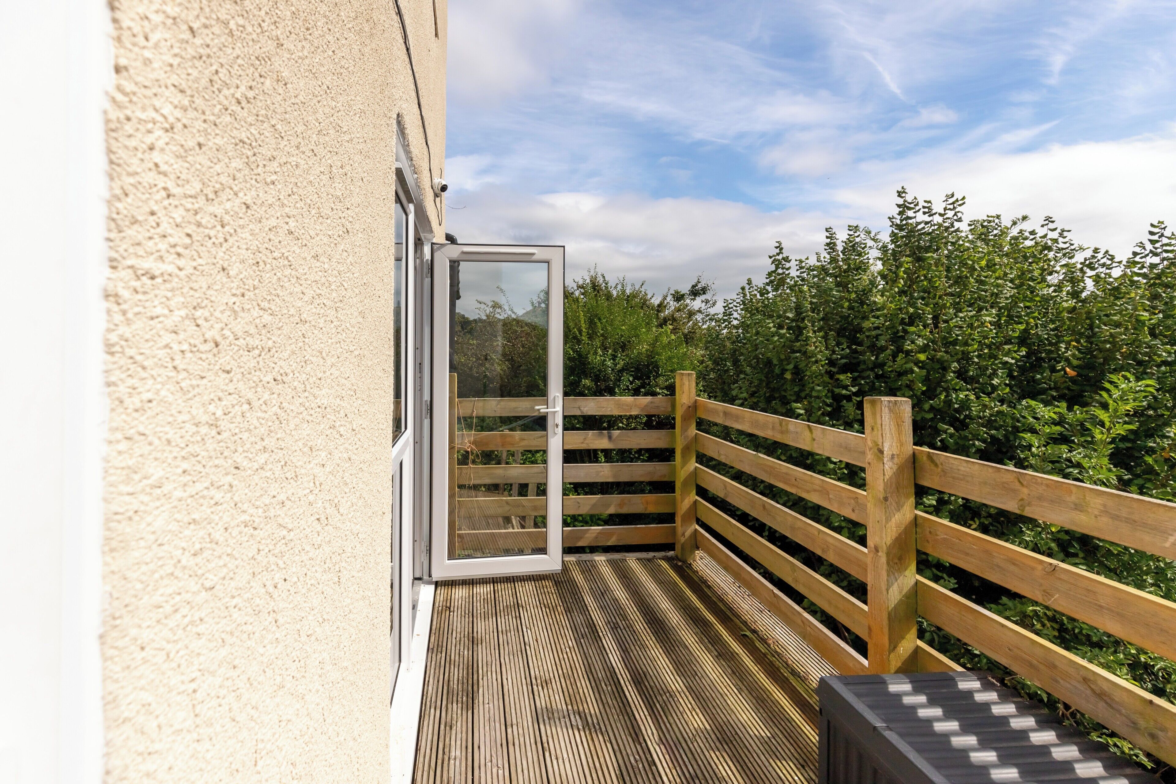 A wooden deck with a glass door leading to a balcony, surrounded by greenery.