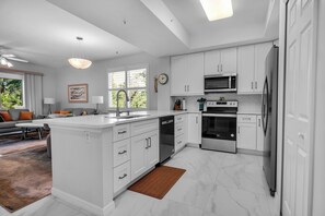 A modern kitchen with white cabinetry, a central island, stainless steel appliances, and a dining area with a sofa and table. - Golf & Beach Shuttle | Estero | 30 Days (Estero)