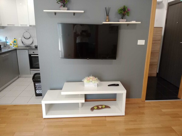 Una sala moderna con un televisor de pantalla plana montado en la pared, un mueble blanco para entretenimiento con un control remoto y una pequeña planta en maceta sobre una estantería.