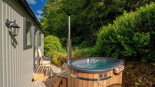 A wooden hot tub with a metal pole, surrounded by a deck and a wooden chair, set against a backdrop of dense greenery.