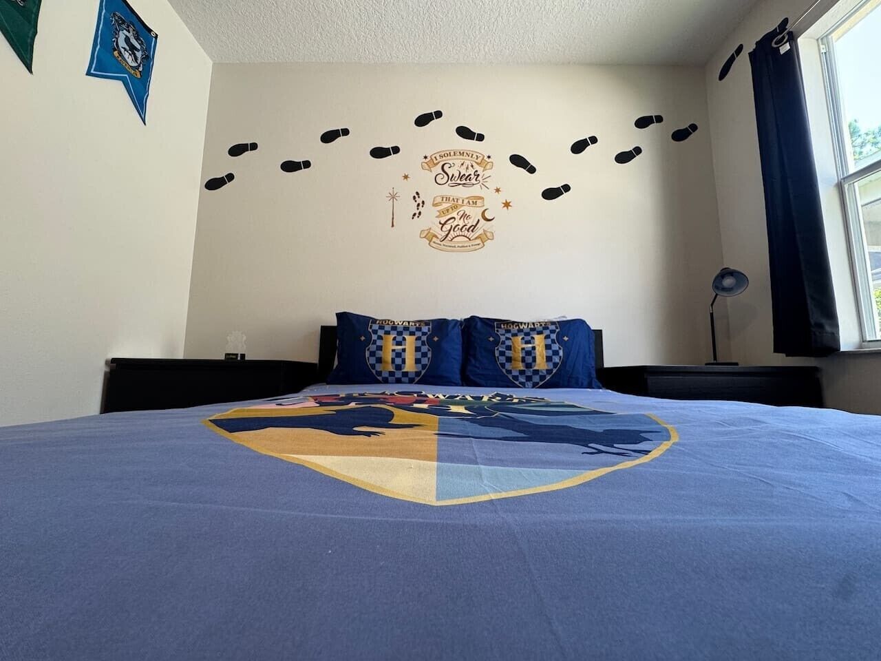 Ett sovrum med en säng som har ett täcke med Hogwarts-motiv, en väggdekor med fotspår och ett citat, och ett fönster med gardiner.
