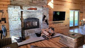 Een chaletinterieur met een stenen openhaard, houten wanden, een aan de muur bevestigde televisie en een gezellige zithoek.