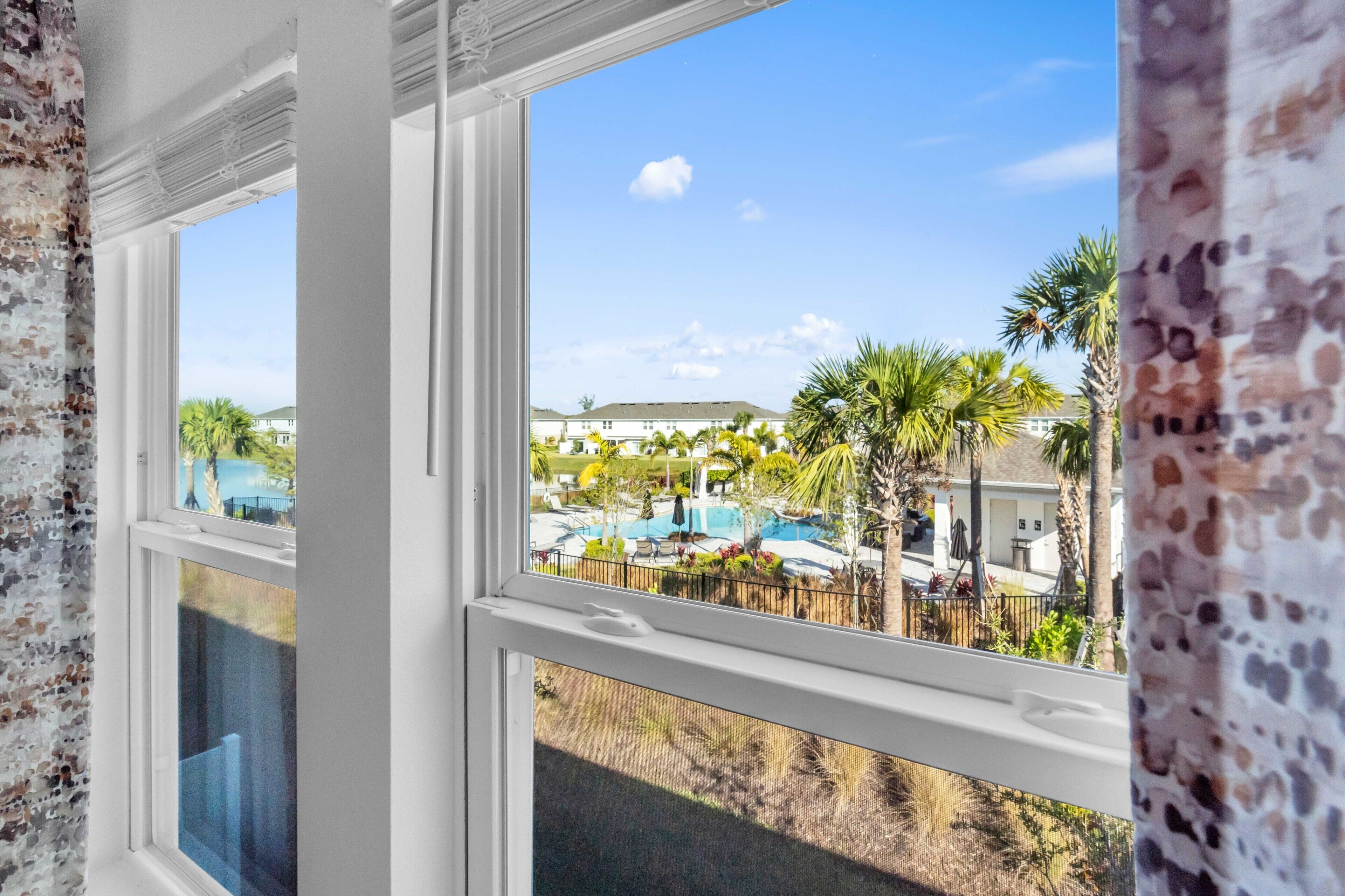 A view through a window with a white frame, a clear blue sky, and a pool area with palm trees and a fence.