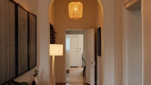 Um corredor com piso de madeira, luminária pendente, uma porta no final e quadros nas paredes.