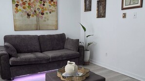 A modern living room with a grey sofa, a coffee table with a decorative item, a ceiling fan, and wall art. - Lovely 2-bedroom house with AC near charming Delaware Bay (Lower Township)