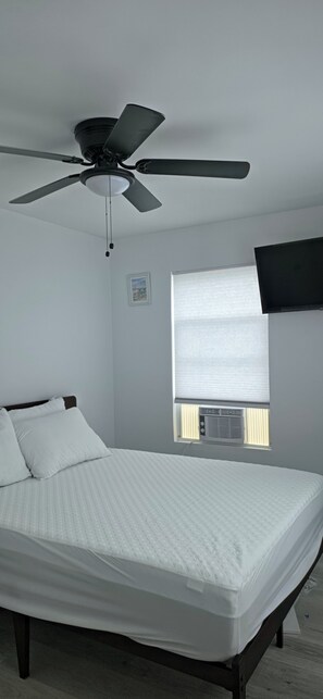 Une chambre à coucher comprenant un lit, un ventilateur de plafond, une fenêtre avec des stores et un téléviseur fixé au mur.