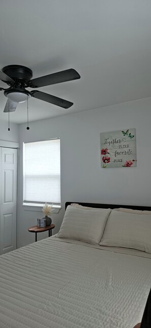 Ein Schlafzimmer mit einem Bett, einem Deckenventilator, einem Fenster mit Jalousien, einem Nachttisch mit einer Vase und einer Wand mit einem Motivationsspruch.