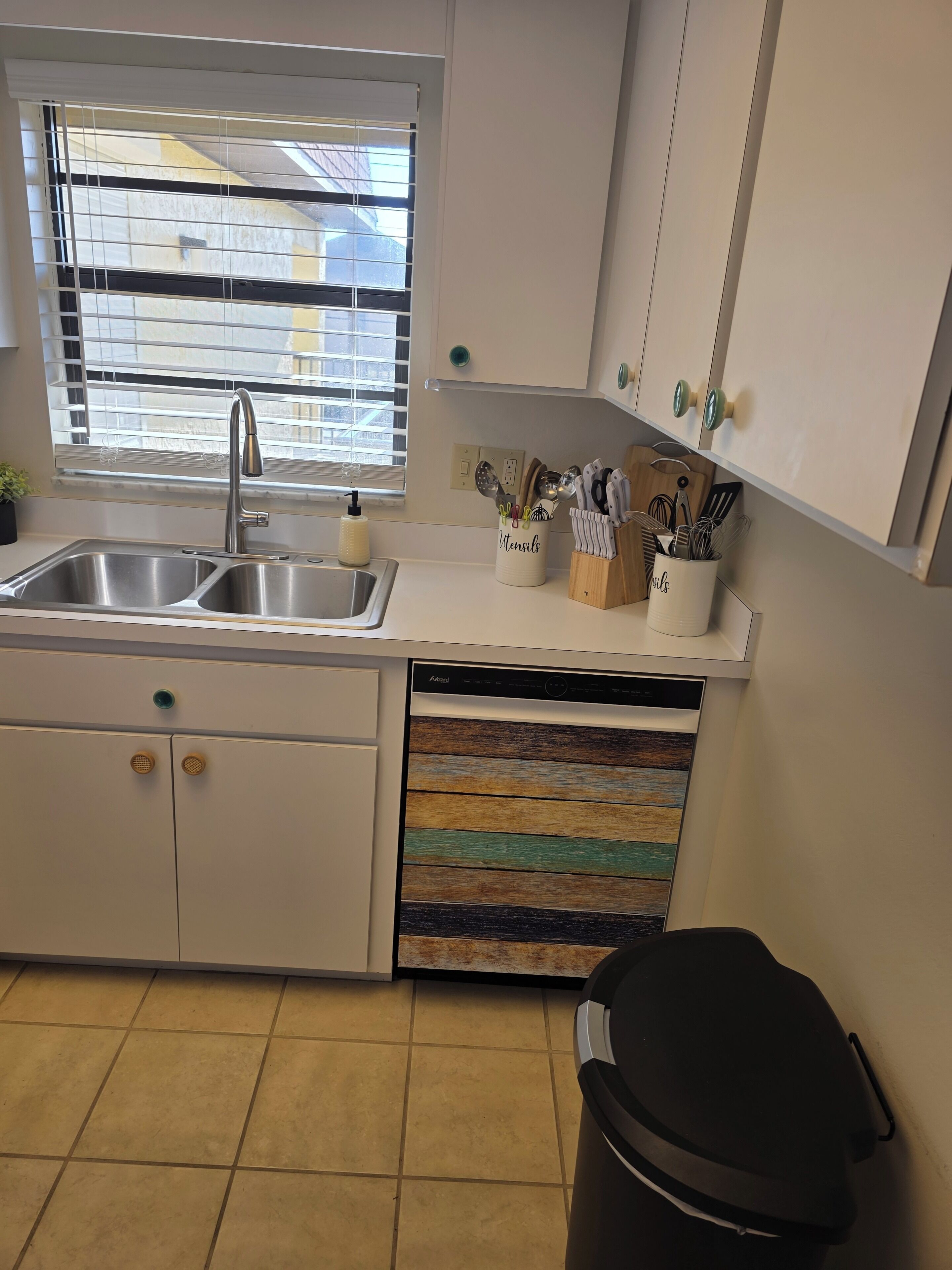 A kitchen with a sink, window, cabinets, and a trash bin.