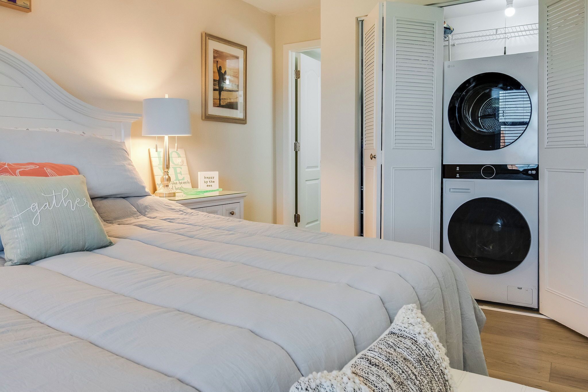 A bedroom with a bed, a nightstand, a lamp, a framed picture, and a wall-mounted washing machine.