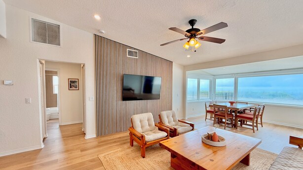 A spacious living room with a wooden coffee table, two armchairs, a TV mounted on a striped wall, and a dining area with a table and chairs. - Direct Oceanfront House – Newly Renovated with Stunning Views-Sleeps 5-2bed2bath (Flagler Beach)