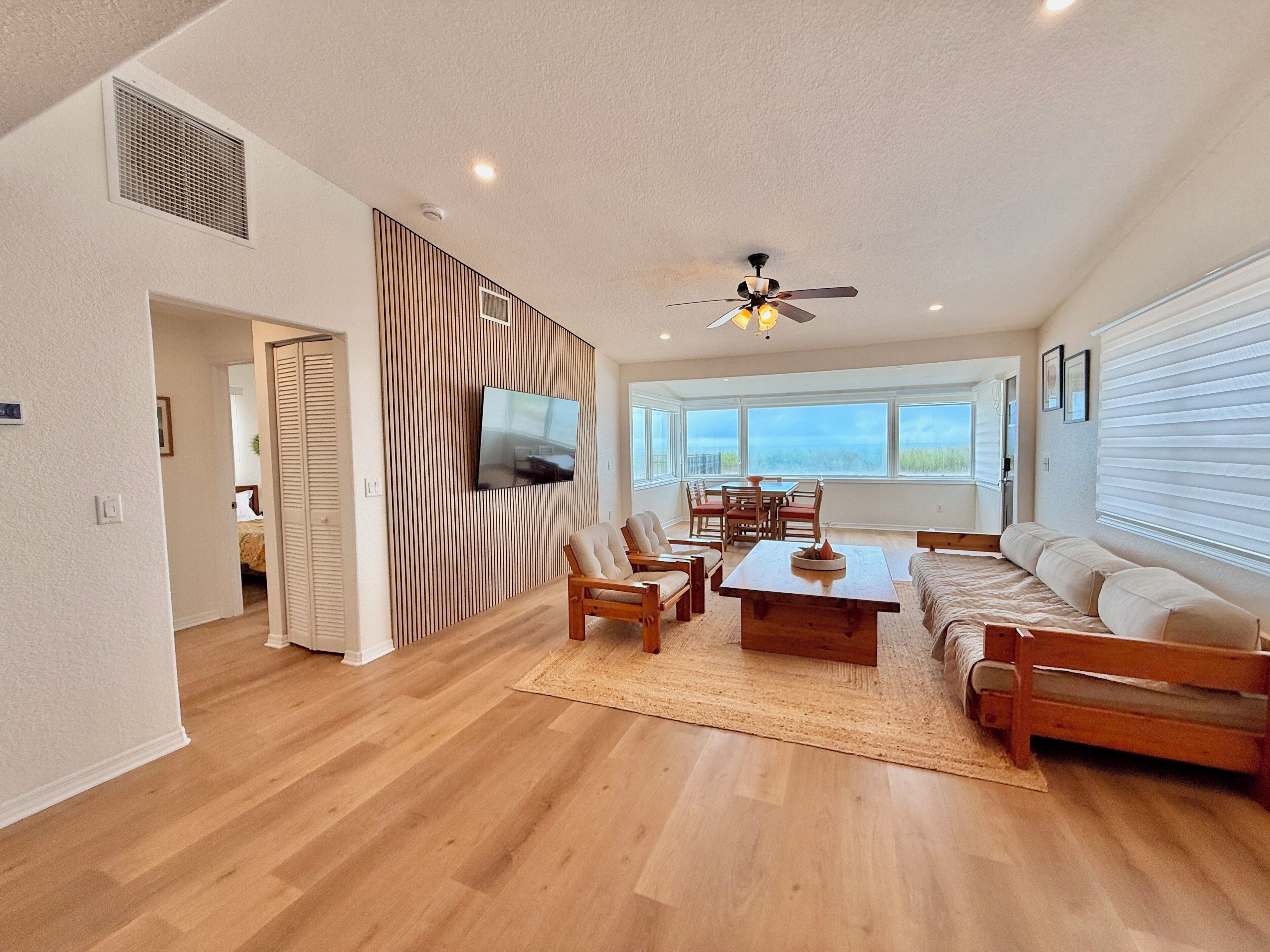 A spacious living room with a wooden floor, a ceiling fan, a flat-screen TV, a dining area with chairs and a table, and large windows offering a view.