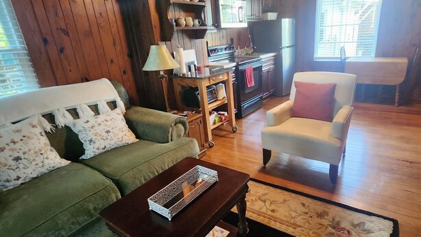 A cozy living room with a green sofa, a white armchair, a wooden coffee table, and a kitchen area with a stove and cabinets.