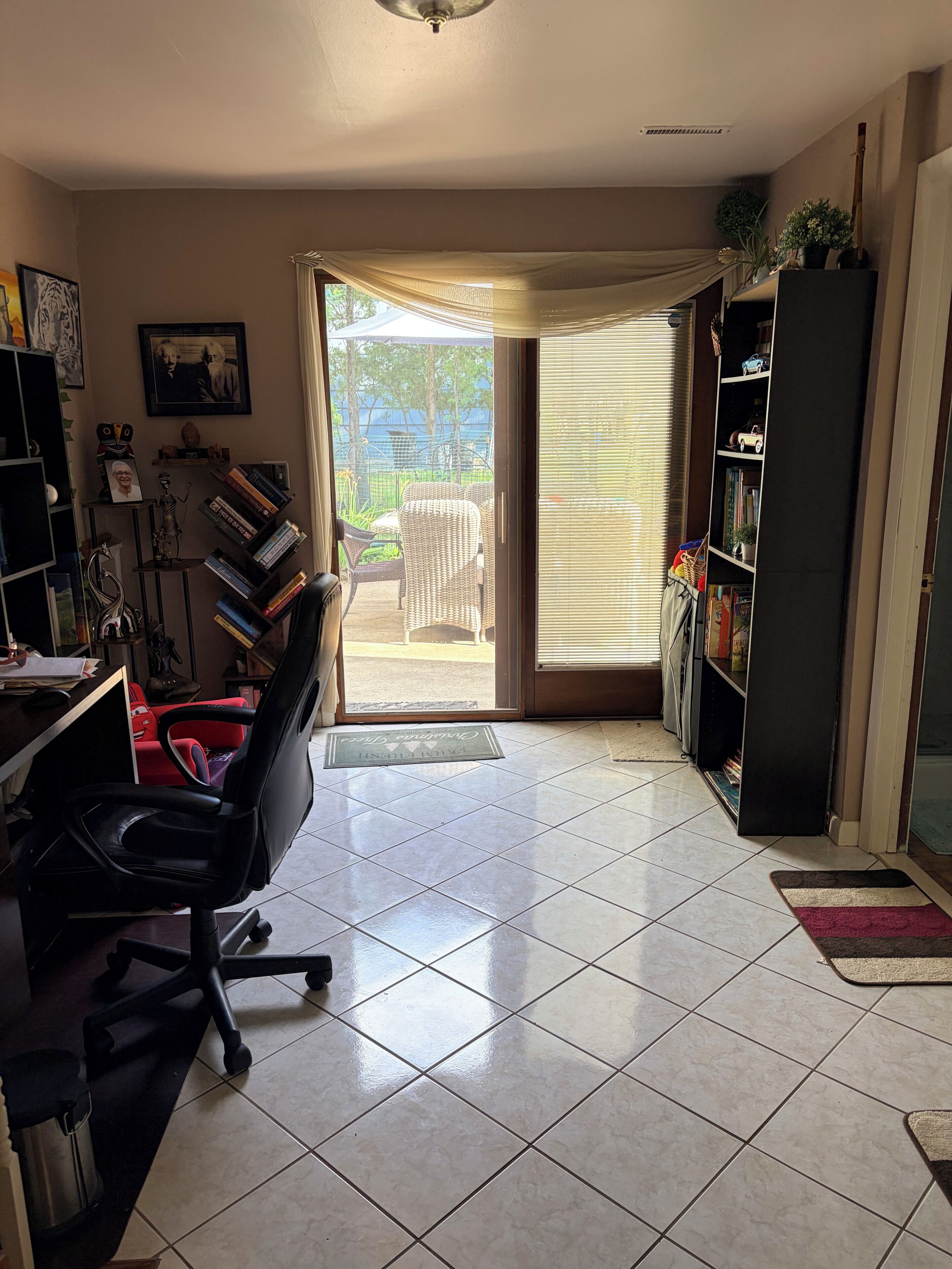 Een kamer met betegelde vloer, een zwarte bureaustoel, een boekenkast, een deur naar buiten en een klein tapijt.